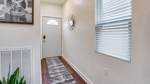 A hallway with a door, a radiator, a plant, a geometric wall decoration, and a window with blinds. - Dog-Friendly Sheboygan Home - 1 Mi to Town! (Sheboygan)