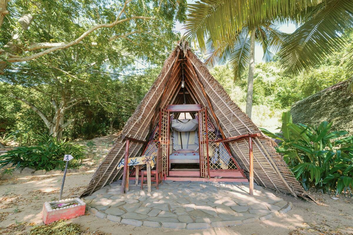 A thatched-roof structure with a bed and a small table, surrounded by lush greenery.