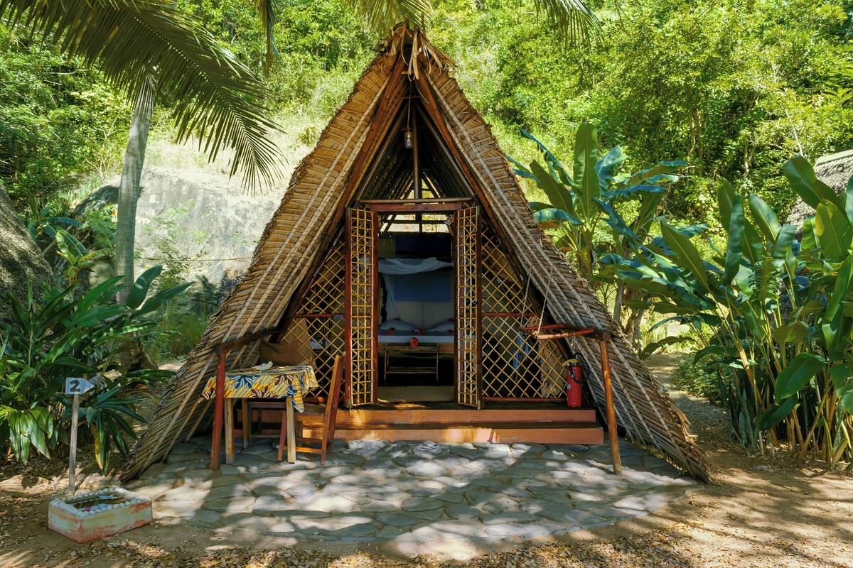 A thatched-roof hut with a bed and a table, surrounded by lush greenery.