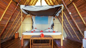 A cozy, rustic bedroom with a wooden floor, a bed with a canopy, and a small table in the center. - Coin Sauvage (Nosy Be)
