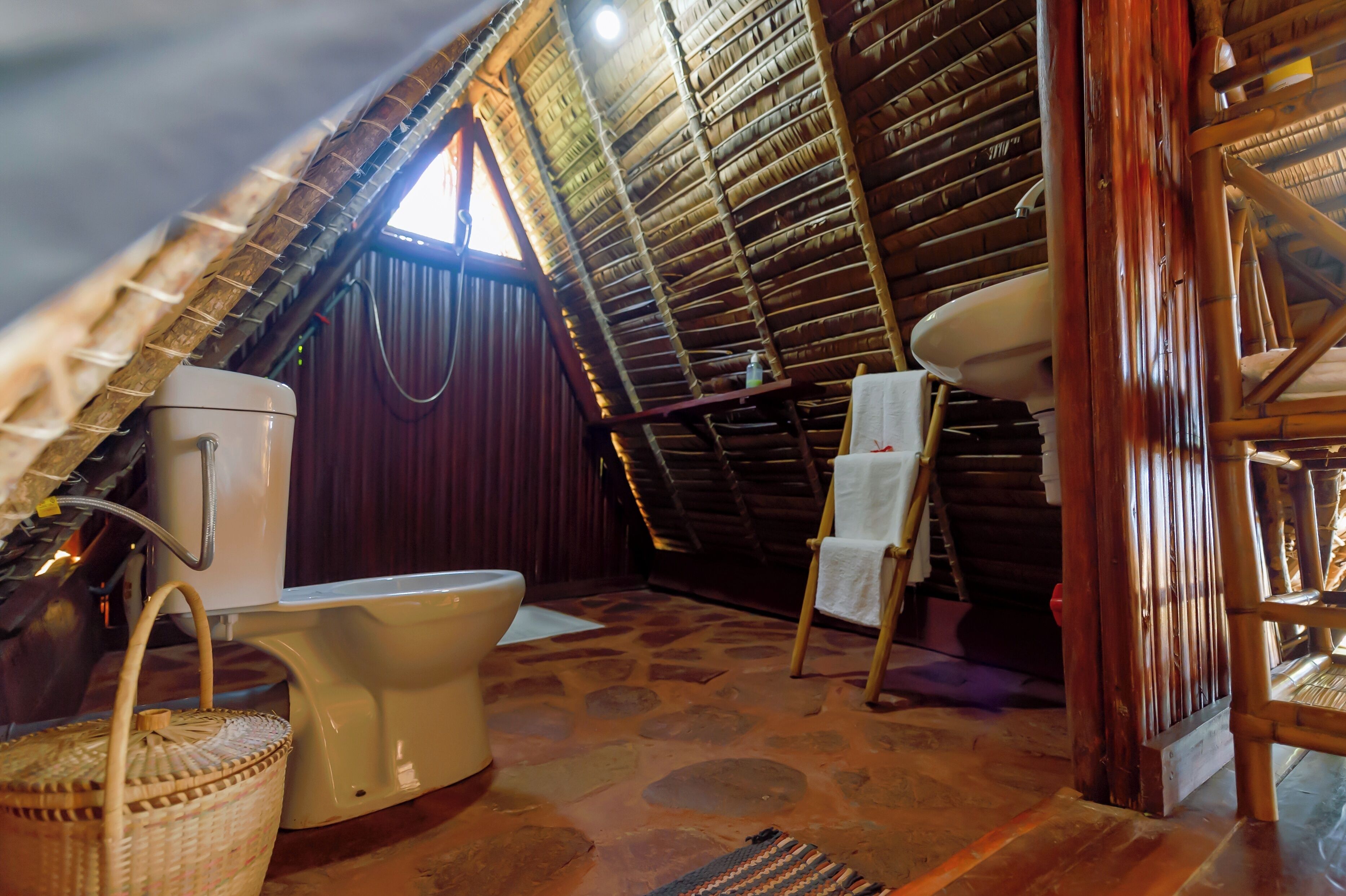 A rustic, wooden interior with a thatched roof, a toilet, a sink, and a chair with a towel.