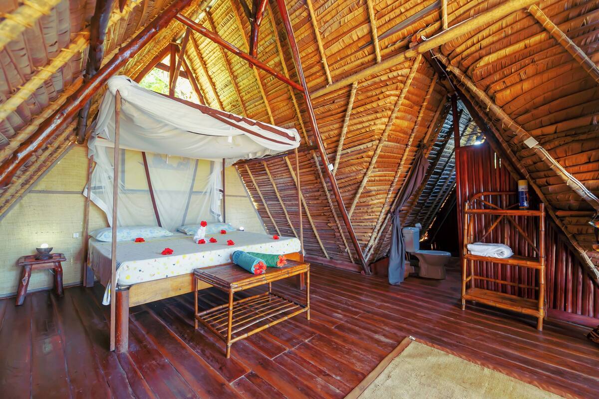 A high-ceilinged, bamboo-framed room with a bed, a small table, and a shelf.