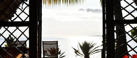 Vista a la playa a través de una ventana de madera con celosías, una silla y una planta.