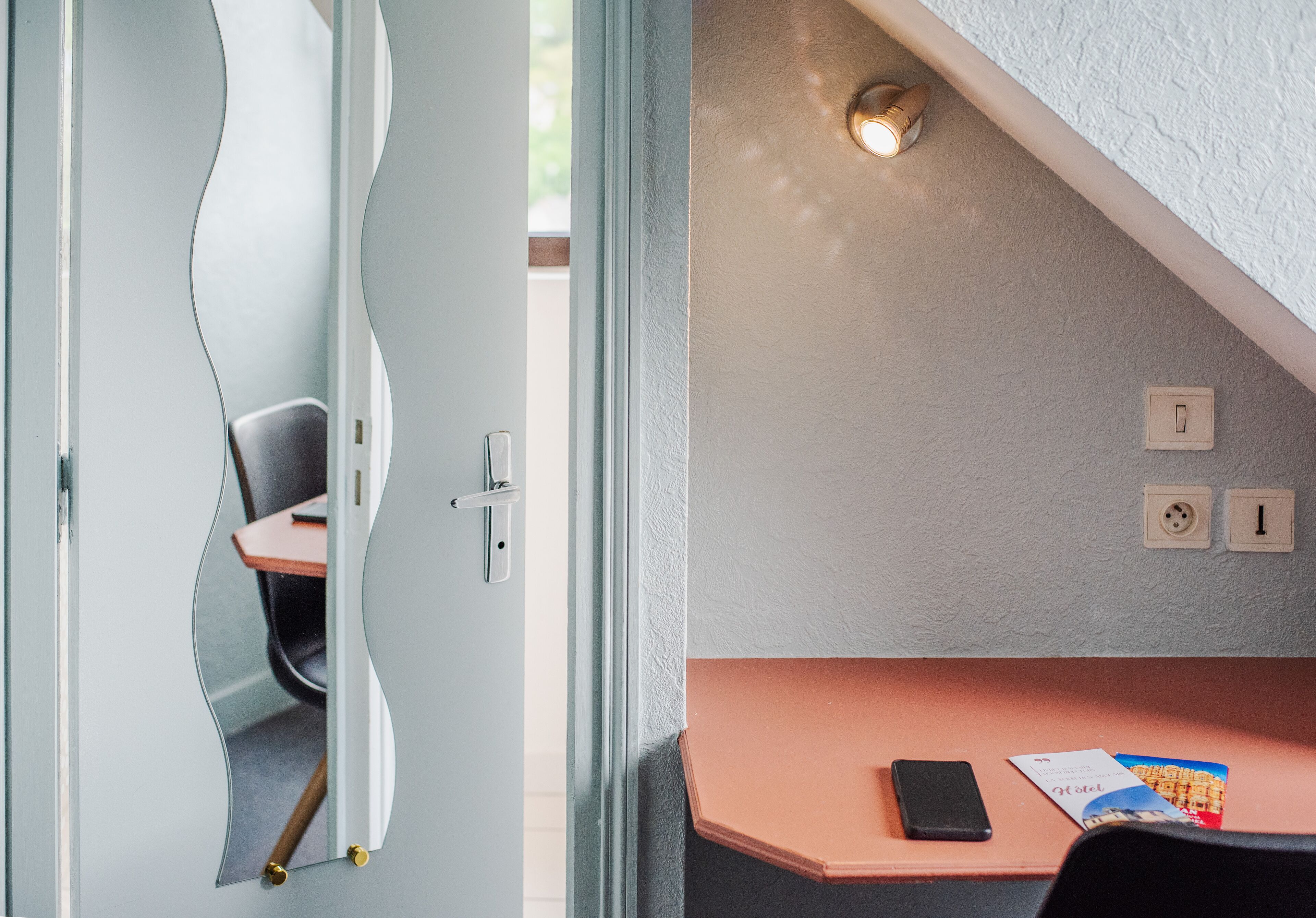 Una habitación moderna con un espejo curvo, un escritorio con teléfono y una revista, y una lámpara en la pared.