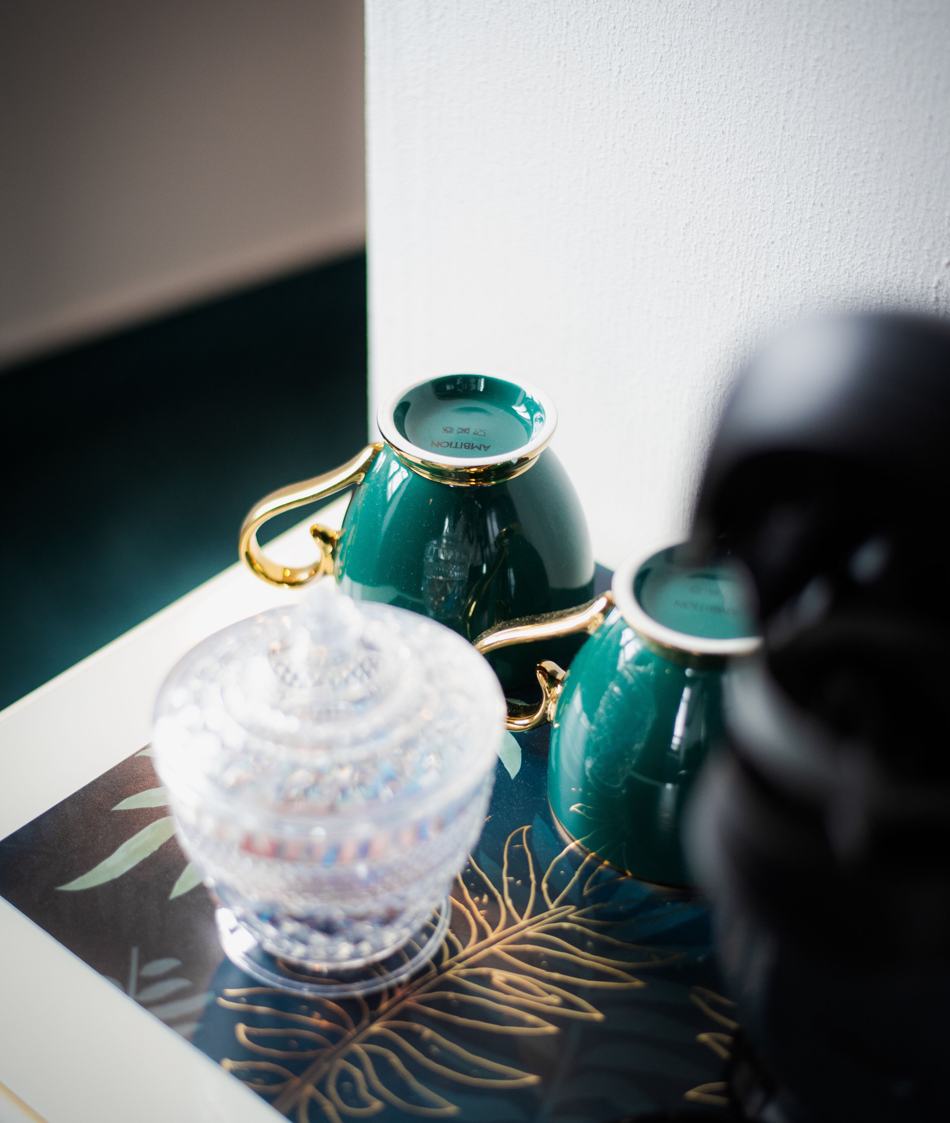 A table with a decorative glass container, two green ceramic cups, and a black object.
