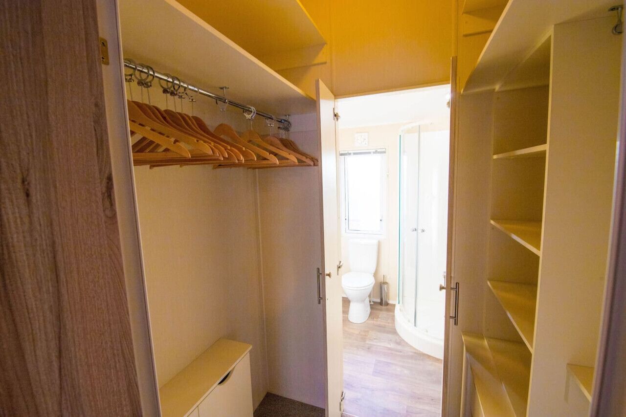 Un dressing accessible sans porte, avec des cintres et des étagères en bois, des toilettes visibles à travers une porte ouverte et un plancher de couleur claire.