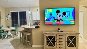A kitchen with a dining area, a TV mounted on the wall displaying a cartoon character, and a wooden cabinet with drawers. - Sunshine 3bdrm pool house near Disney (Kissimmee)