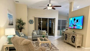 A living room with a sofa, armchair, coffee table, TV, and a sliding door leading to another room. - Sunshine 3bdrm pool house near Disney (Kissimmee)