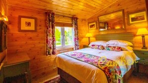 A wooden cabin bedroom with a bed, two bedside lamps, a window with curtains, and framed pictures on the walls. - Rainbow Petty - Pine lodge - Kent countryside (Brookland)