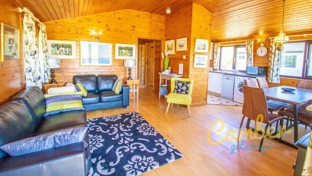 L’intérieur chaleureux d’un chalet avec un plafond en bois, un espace de vie comprenant un canapé et des fauteuils, une table à manger avec des chaises, et un coin cuisine.