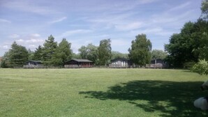 Un vaste champ herbeux avec plusieurs constructions en bois, peut-être des cabanes ou des dépendances, entouré d’arbres.