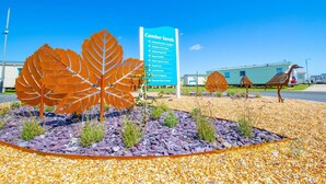 Ein Schild mit der Aufschrift 'Camber Sands' und Auflistung der Annehmlichkeiten, umgeben von skulpturalen, blattformigen Elementen und Außenmöbeln.