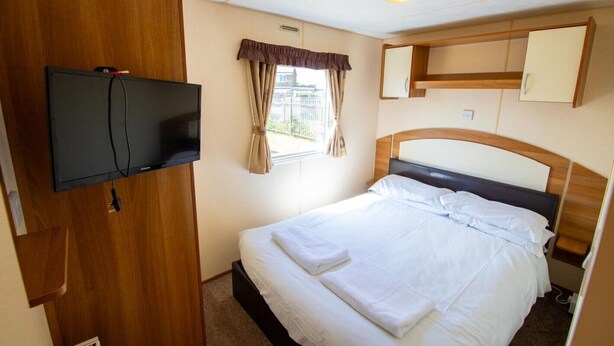 A bedroom with a bed, a television mounted on a wooden cabinet, a window with curtains, and a headboard.
