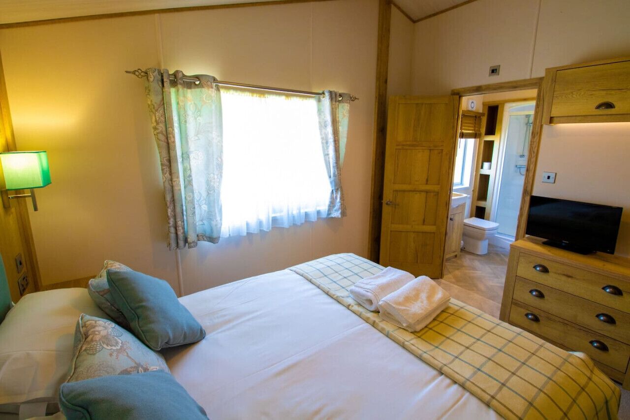 A bedroom with a bed, pillows, a television, a window with curtains, a bathroom with a toilet, and a wooden cabinet.