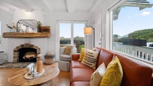 A cozy living room with a stone fireplace, a leather sofa, and a large window overlooking a scenic landscape. - #StayInMyDistrict SurfsideHaven (Rockaway Beach)