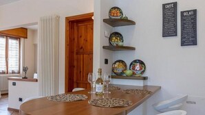 A dining area with a wooden table, white chairs, and a bottle of wine. There are wall-mounted shelves with plates and a menu board.