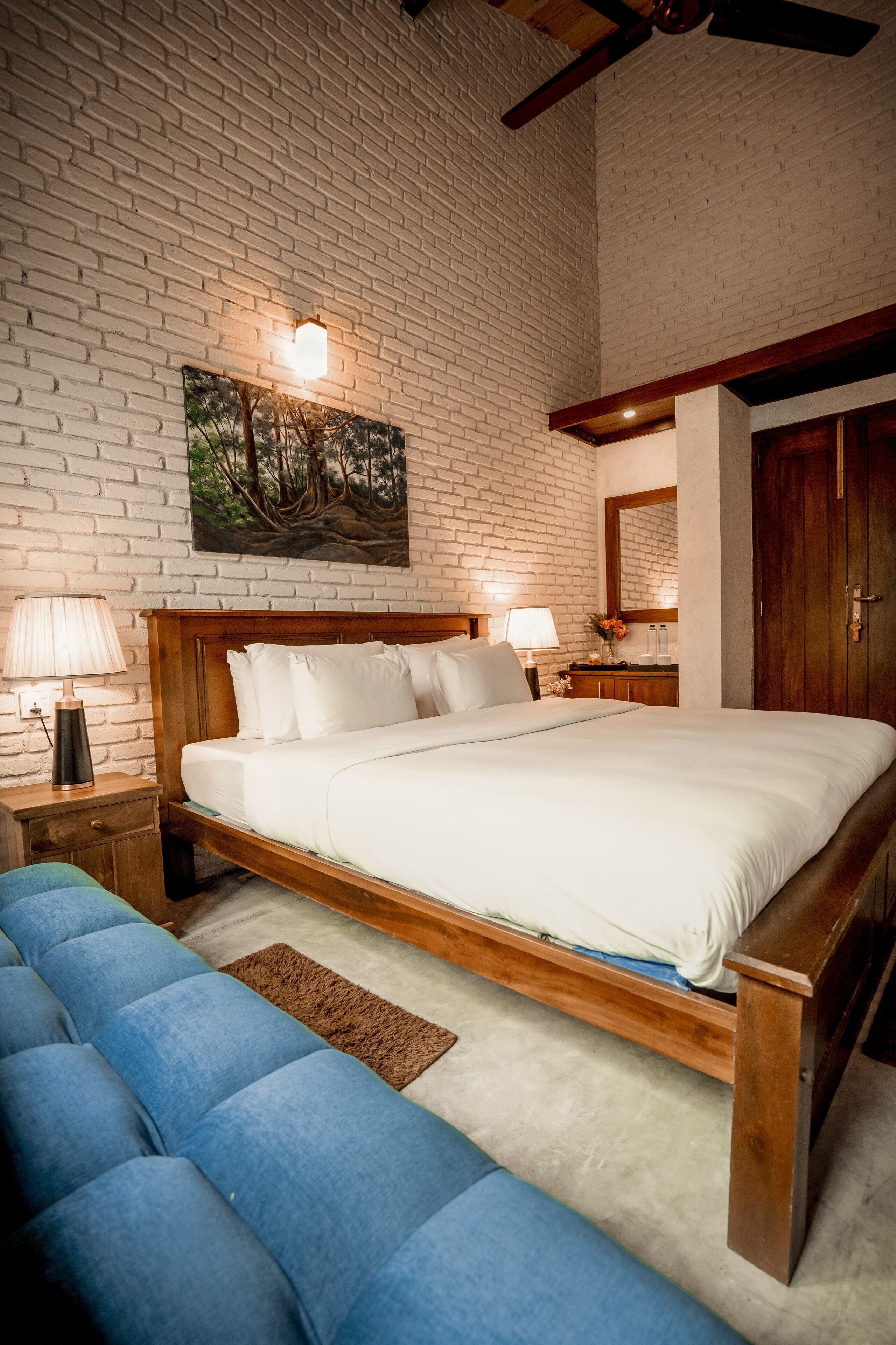 Un dormitorio con cama de madera, ropa de cama blanca, sofá azul, pared de ladrillo y ventilador de techo.