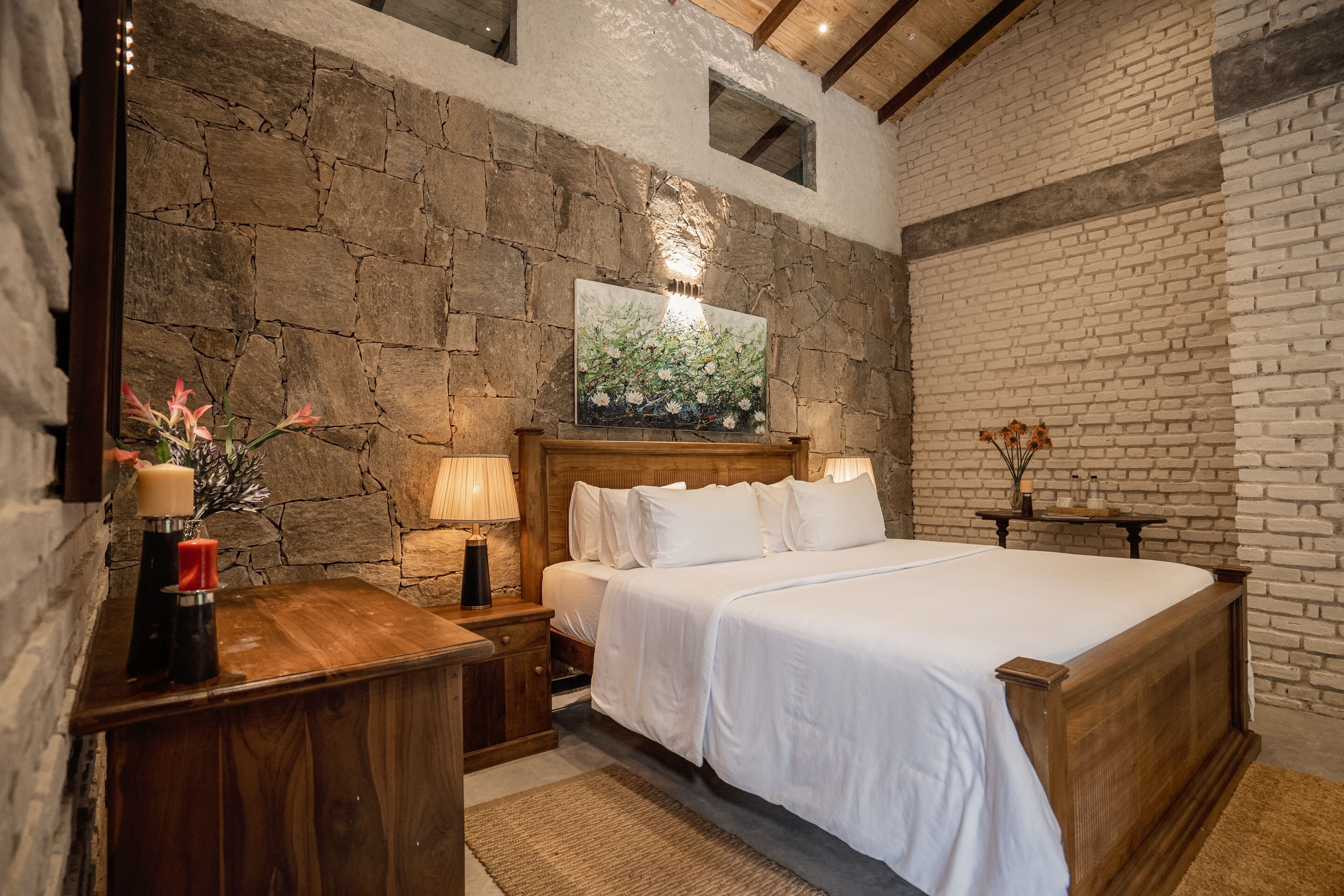 A bedroom with a stone wall, a wooden bed, a nightstand, a lamp, and a painting above the bed.