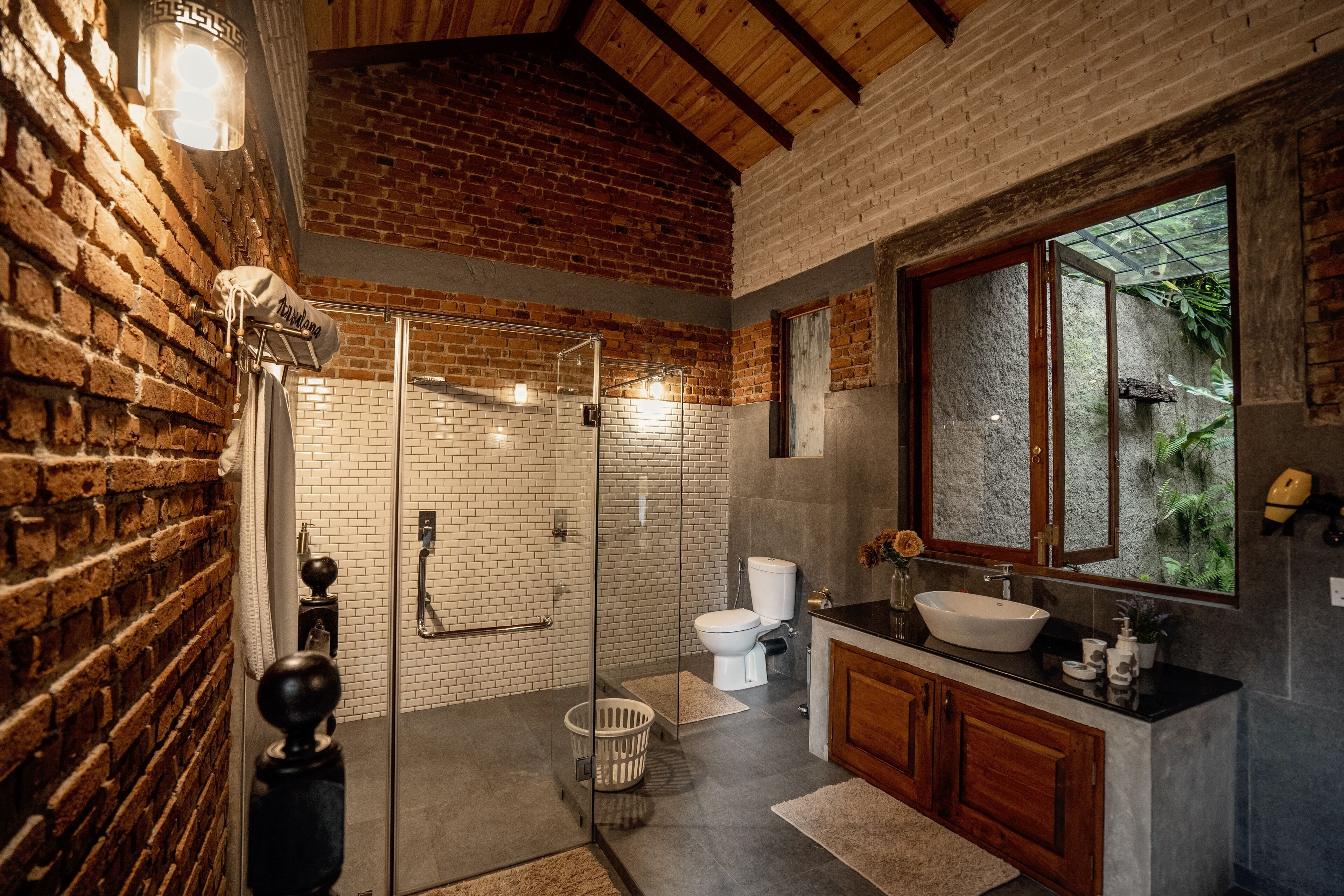 Un baño con pared de ladrillo, cabina de ducha de vidrio, inodoro blanco y tocador de madera con lavabo.