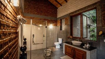 Un baño con pared de ladrillo, cabina de ducha de vidrio, inodoro blanco y tocador de madera con lavabo.