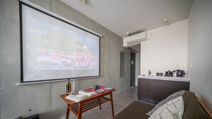 Une pièce moderne avec un grand écran de projection, une table en bois sur laquelle sont posés des livres et une télécommande, un canapé recouvert d’un plaid à motifs, et, au fond, une petite cuisine.