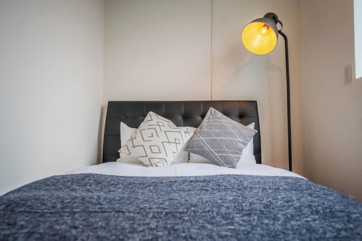 A bed with a dark headboard, white bedding, and patterned pillows. A floor lamp with a yellow shade is positioned to the right of the bed.