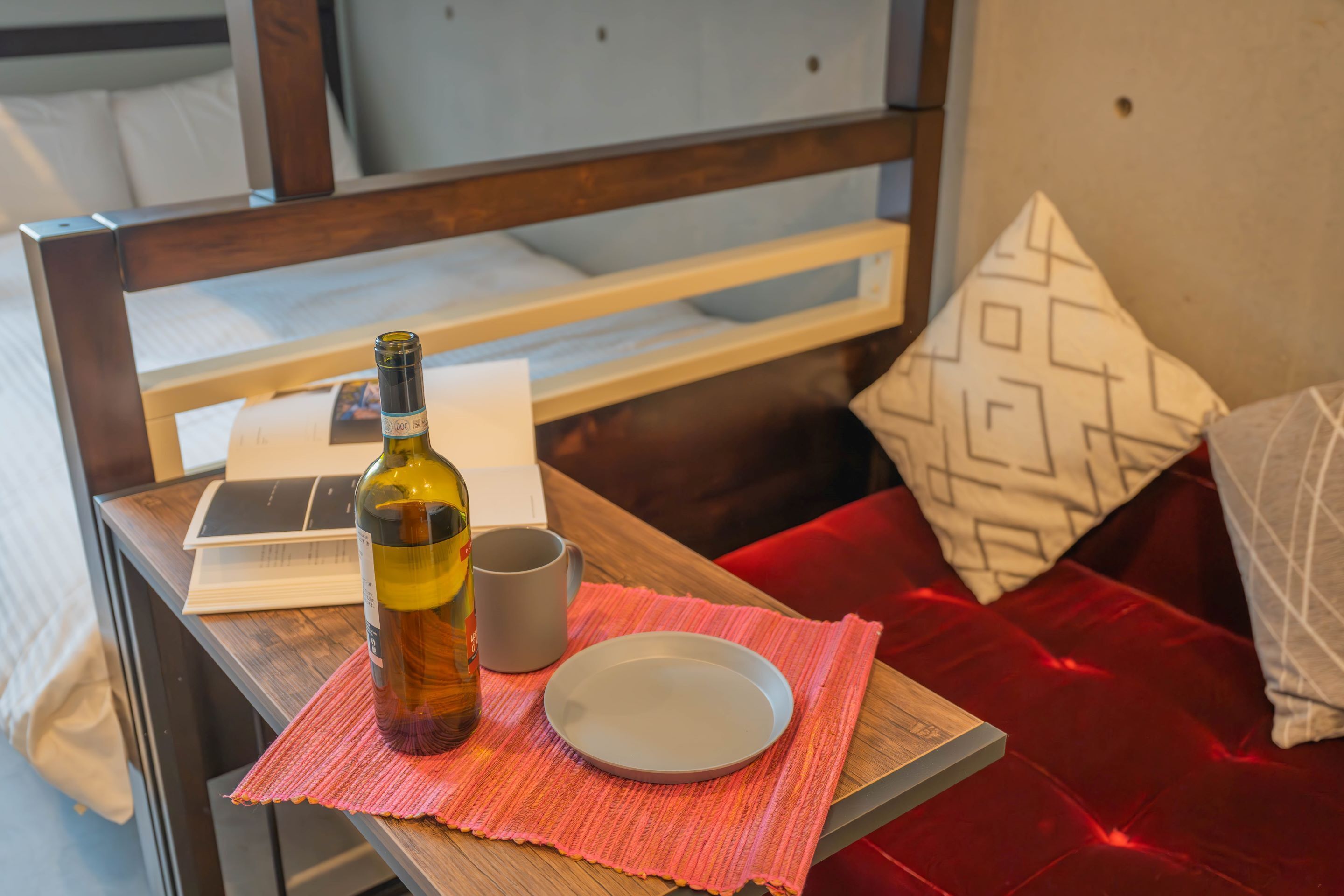 A wooden table with a bottle of wine, a gray mug, and a white plate on a red placemat.