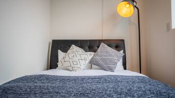 A neatly made bed with a dark headboard, multiple patterned pillows, and a blue blanket. A floor lamp with a yellow light is positioned to the right.