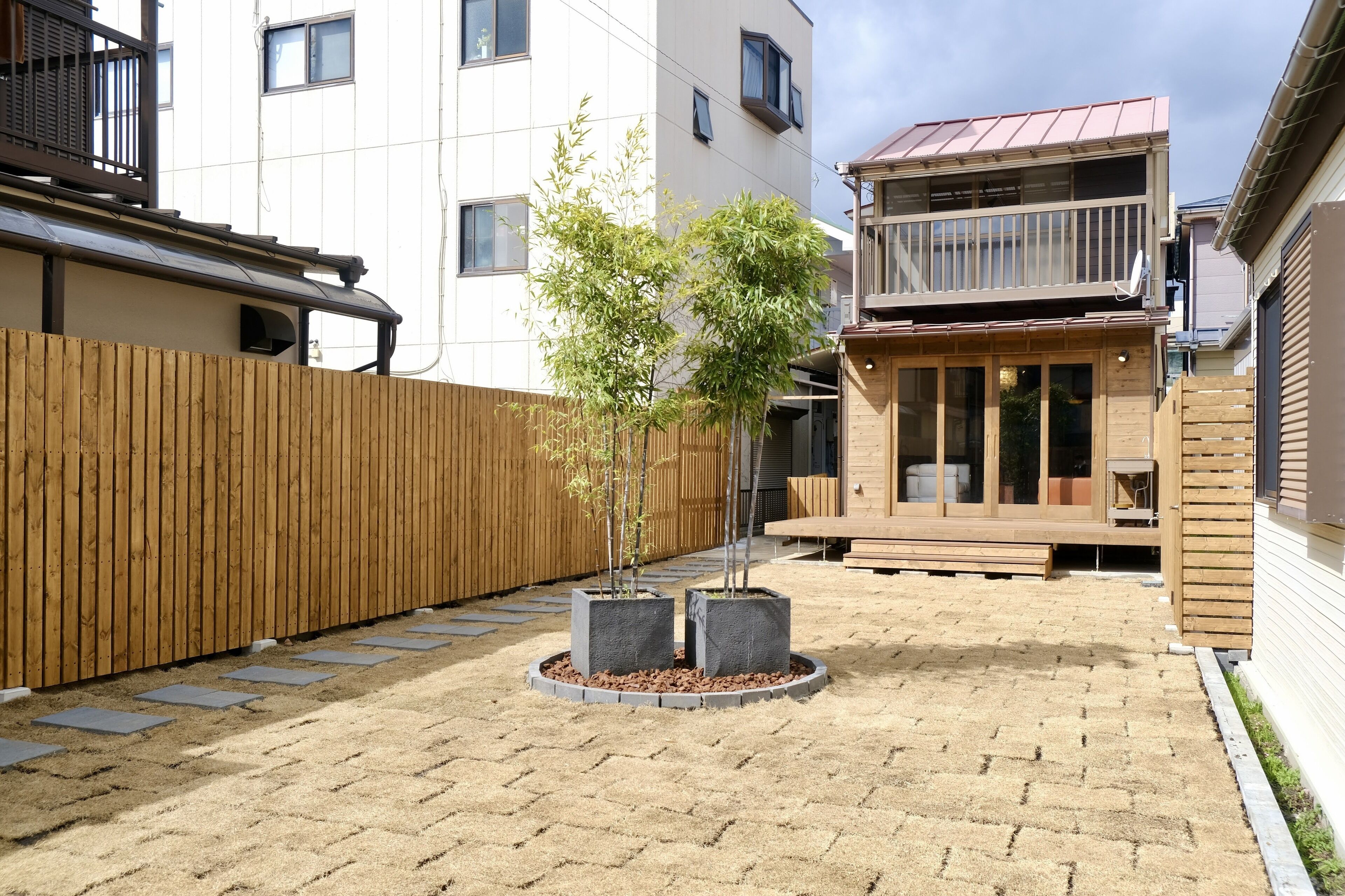 A courtyard with a wooden fence, two potted bamboo trees, and a brick pathway.
