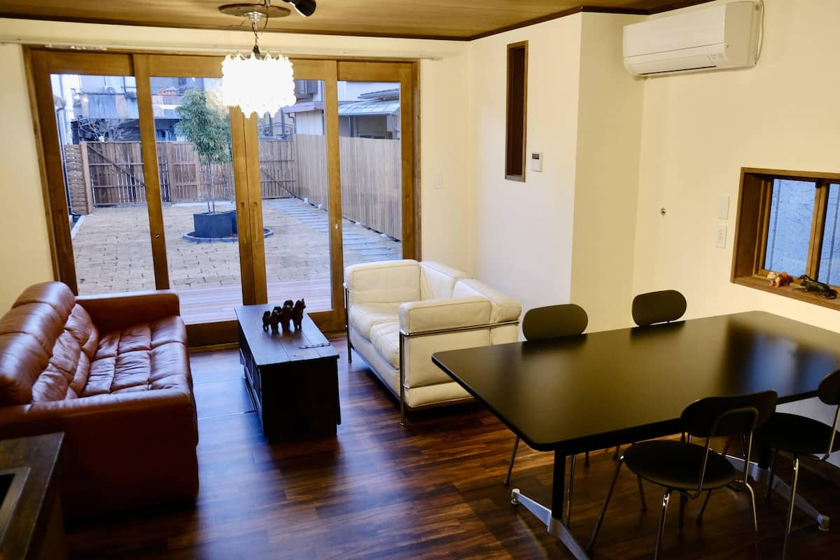 Uma sala de estar com um sofá de couro marrom, um sofá branco, uma mesa de jantar com cadeiras e piso de madeira.
