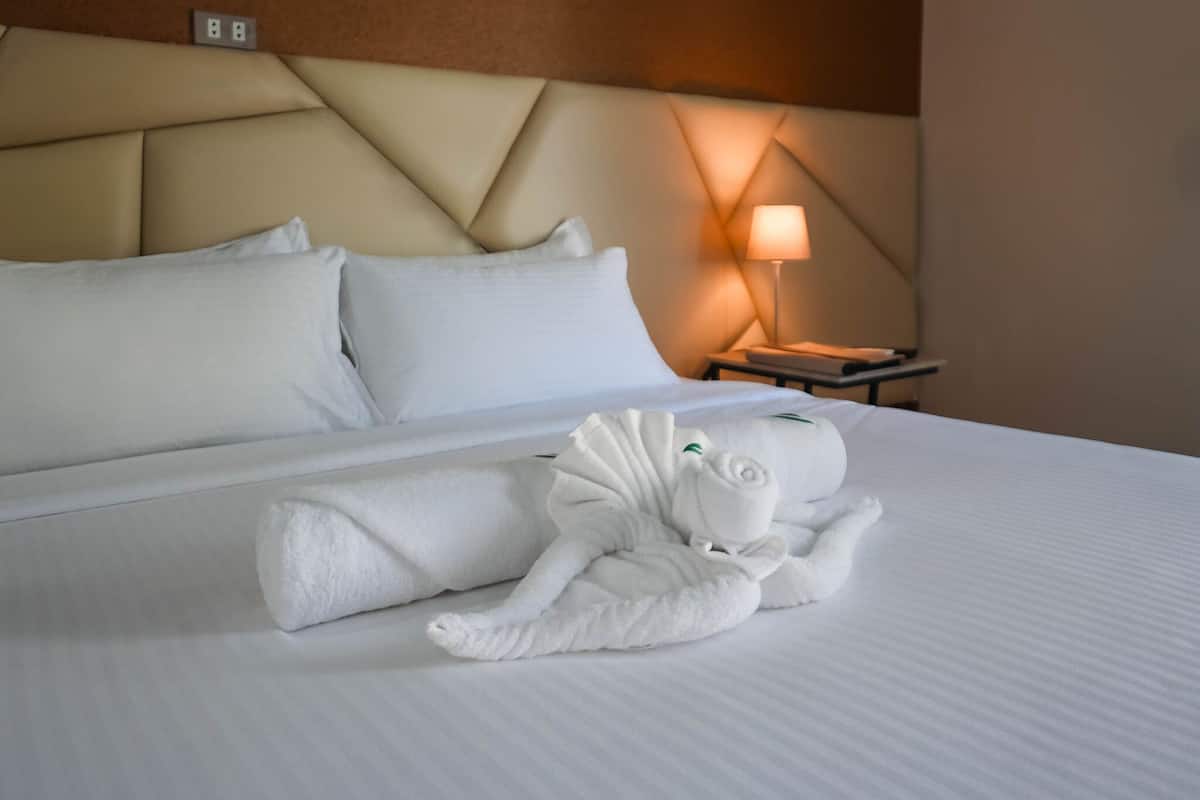 Une chambre d’hôtel avec un lit soigneusement fait, une tête de lit, une table de chevet avec une lampe, et une serviette disposée avec art sur le lit.