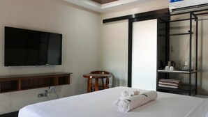 A hotel room with a bed, a TV mounted on the wall, a wooden chair, a shelf with toiletries, and a towel on the bed.