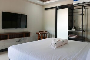 Una habitación de hotel con una cama, un televisor montado en la pared, una silla de madera, un estante con artículos de aseo y una toalla sobre la cama.