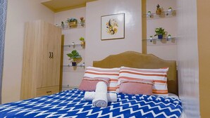 A bedroom with a bed, striped pillows, a rolled towel, a wooden wardrobe, and shelves with plants. - MESAVERTE STUDIO UNIT (Cagayan de Oro)