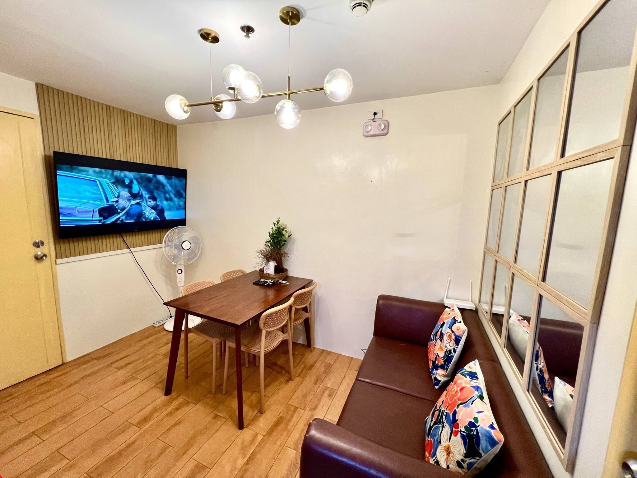 A modern living room with a wooden dining table, a sofa with floral cushions, a TV mounted on the wall, and a ceiling light fixture.
