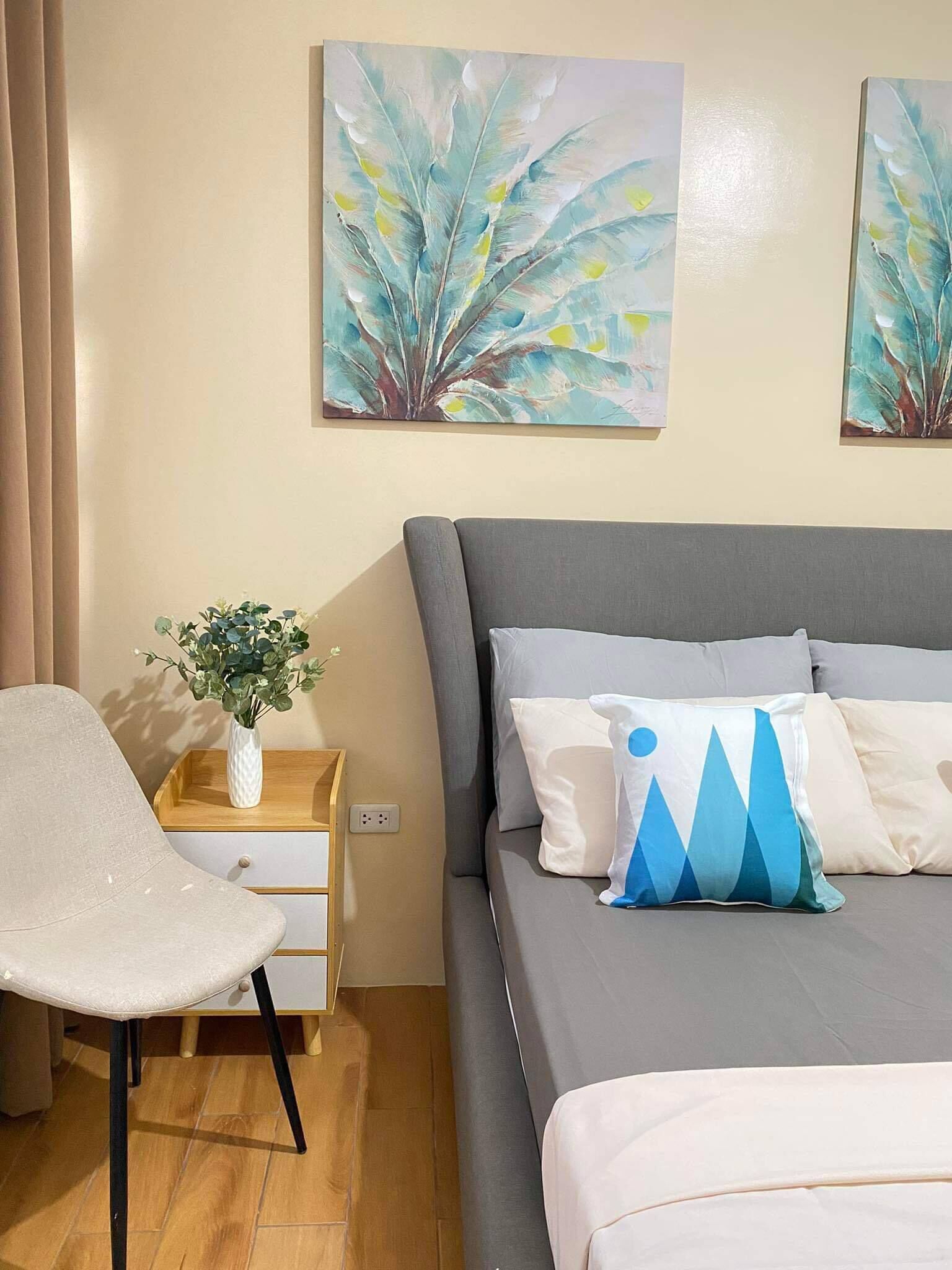 A modern bedroom with a grey bed, white and blue pillows, a white chair, a wooden nightstand with a vase of green plants, and two abstract paintings on the wall.