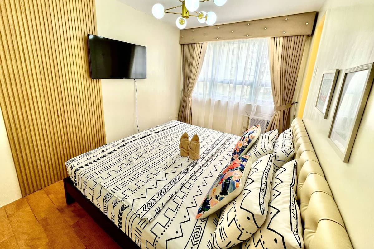 Ein modernes Hotelzimmer mit einem großen Bett, einem Flachbildfernseher, einer Deckenleuchte und einem Fenster mit Vorhängen.