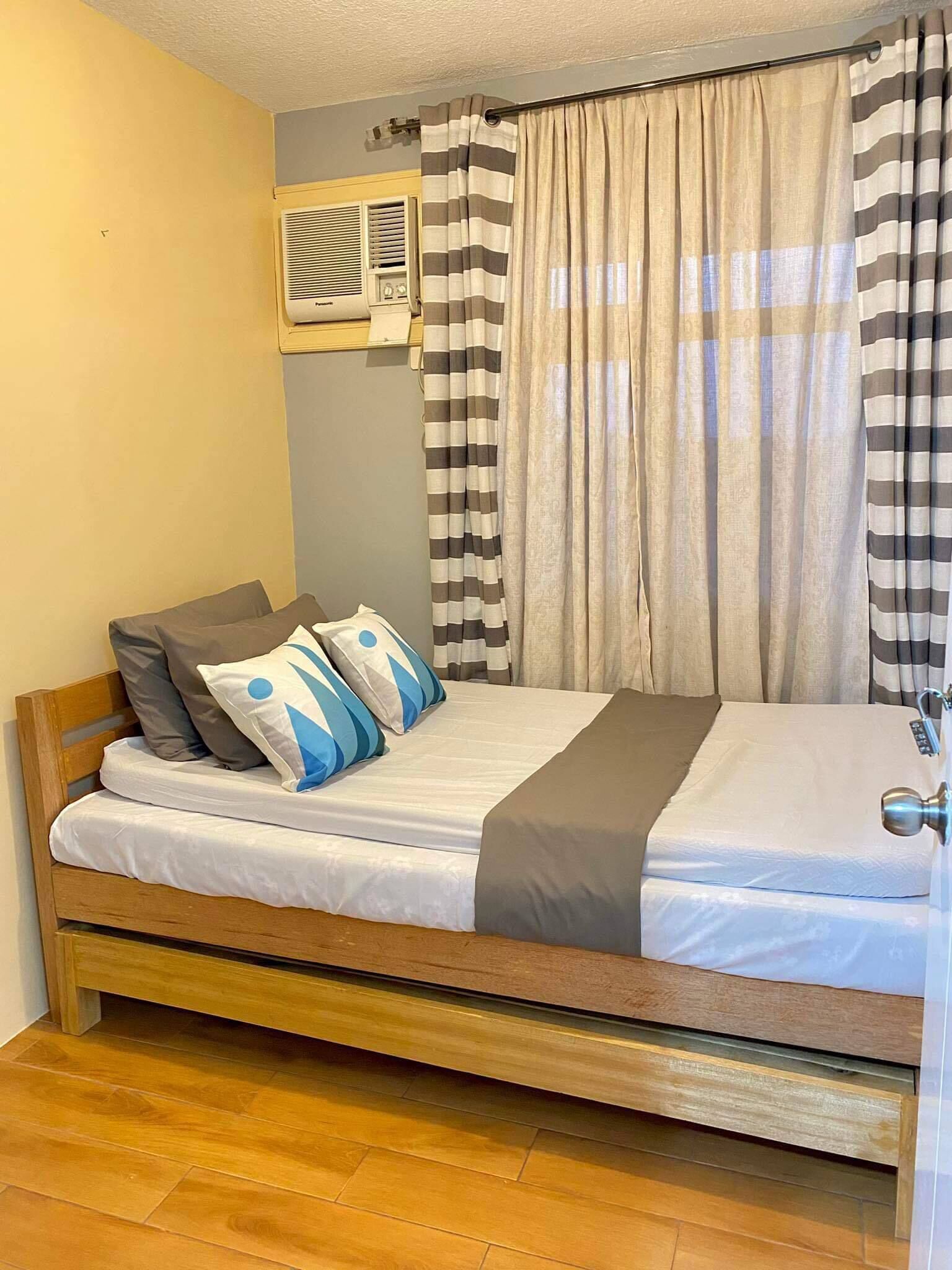 A single bed with a wooden frame, a white mattress, and blue and gray pillows. There is a window with beige curtains and a wall-mounted air conditioning unit.