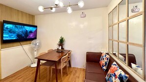 A modern living room with a wooden dining table, a brown sofa with floral cushions, a TV mounted on the wall, and a ceiling light fixture.