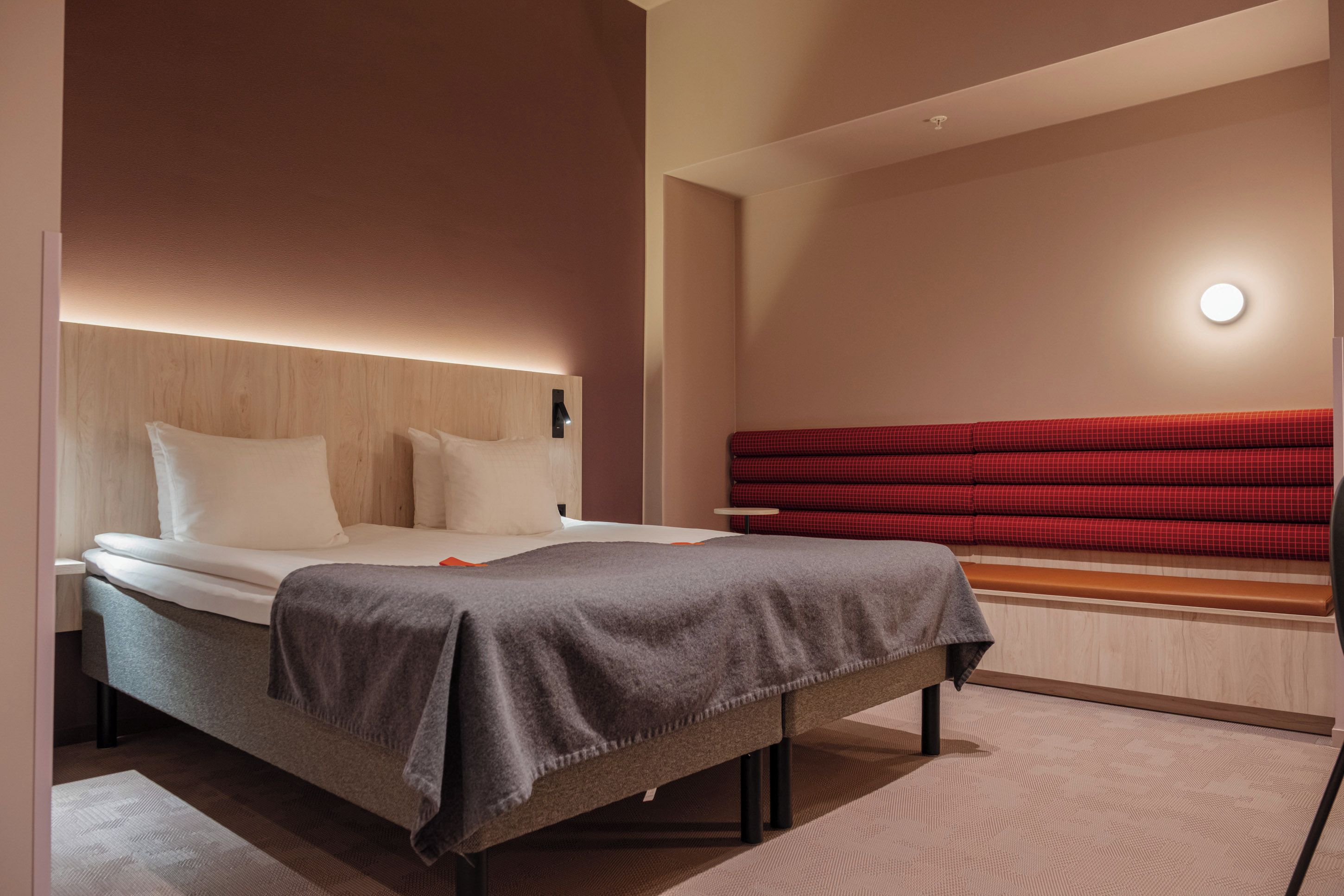 Une chambre d’hôtel moderne avec un lit, un banc recouvert d’un tissu rouge et un luminaire fixé au mur.