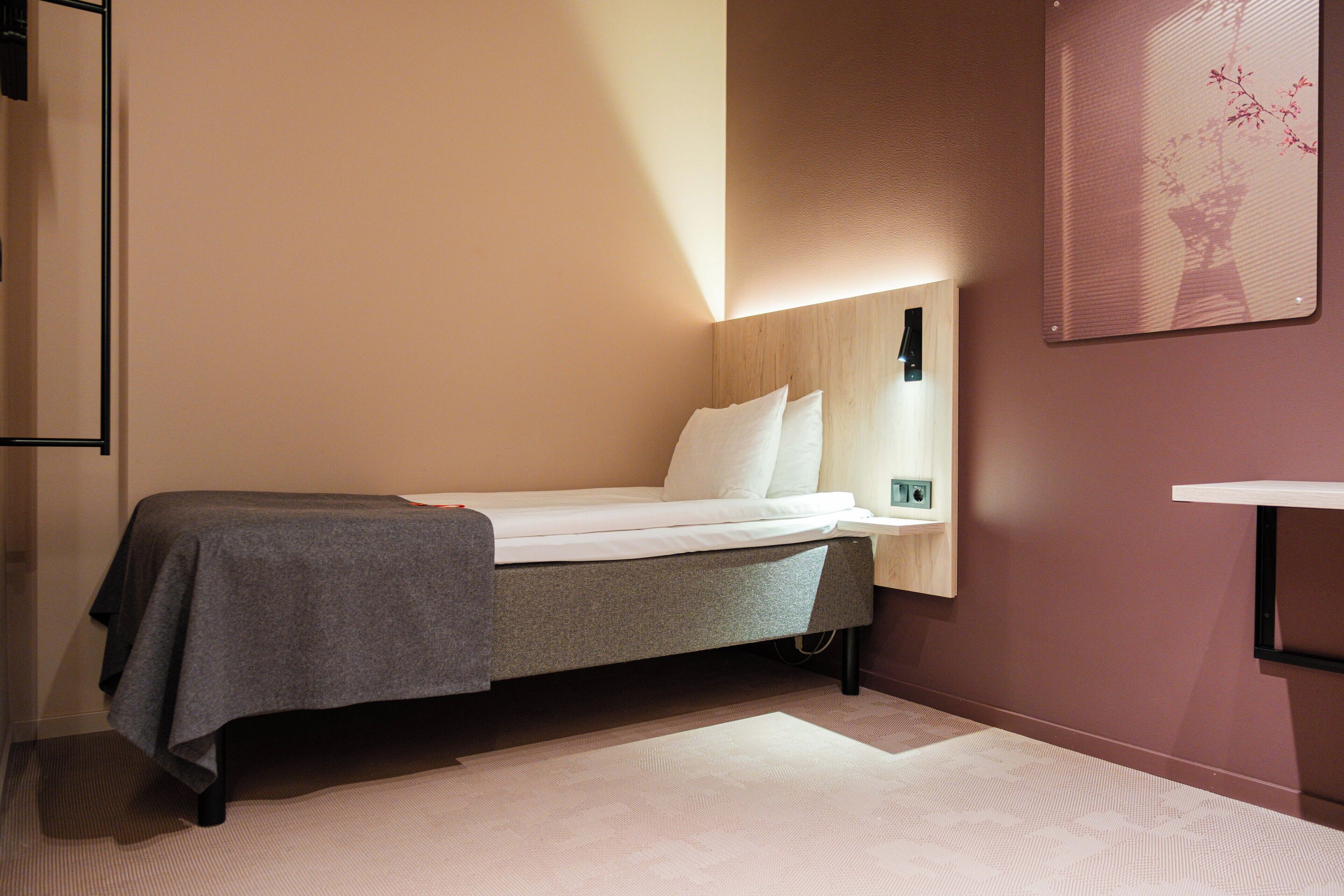 A single bed with a grey blanket, a bedside table with a lamp, and a wall-mounted shelf.