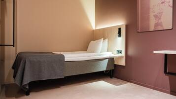 A single bed with a grey blanket, a bedside table with a lamp, and a wall-mounted shelf.