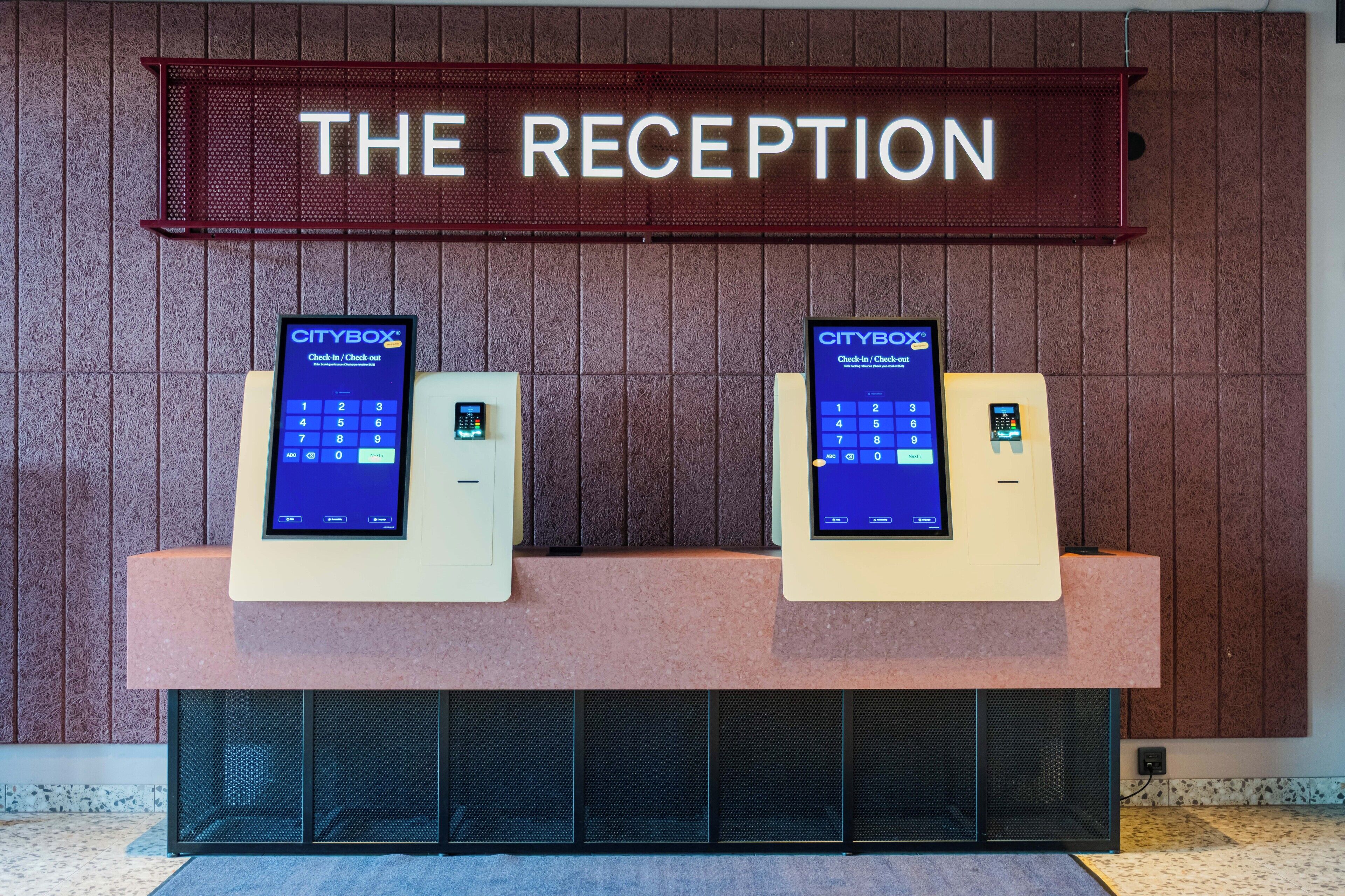 A reception area with two CityBox kiosks for check-in and check-out, a sign above reading 'THE RECEPTION', and a textured wall in the background.