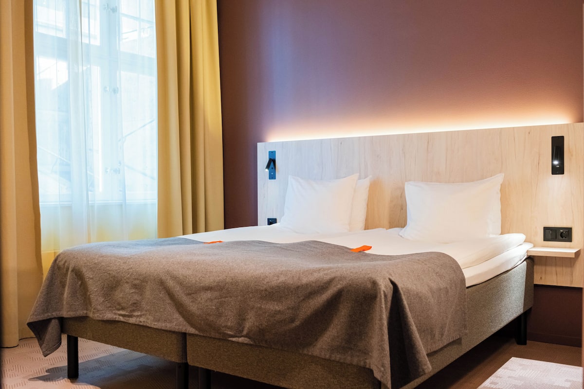 A hotel room with a bed, a nightstand, a wall-mounted light, and a window with curtains.