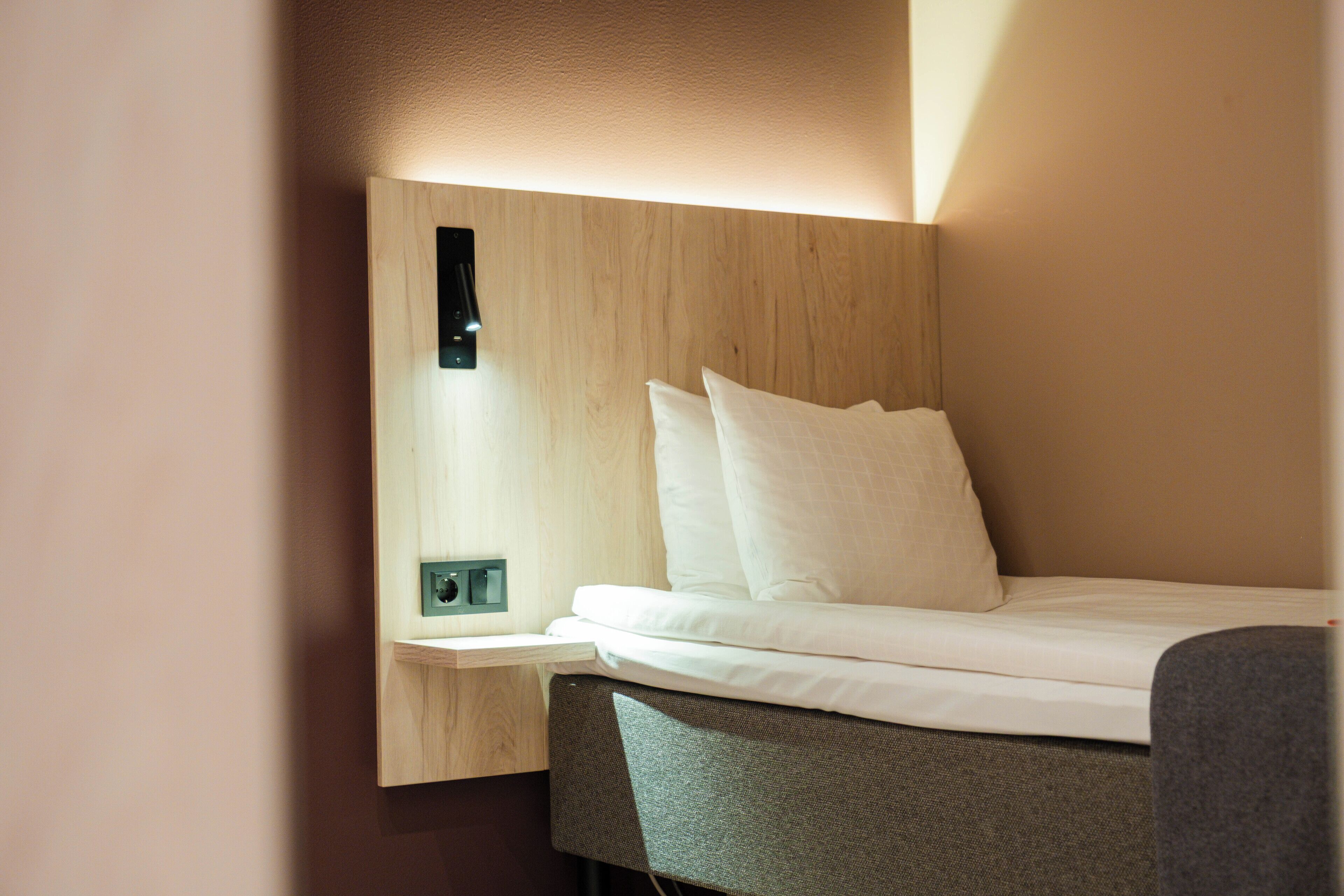 A modern hotel room with a neatly made bed, a bedside table, and a wall-mounted light fixture.
