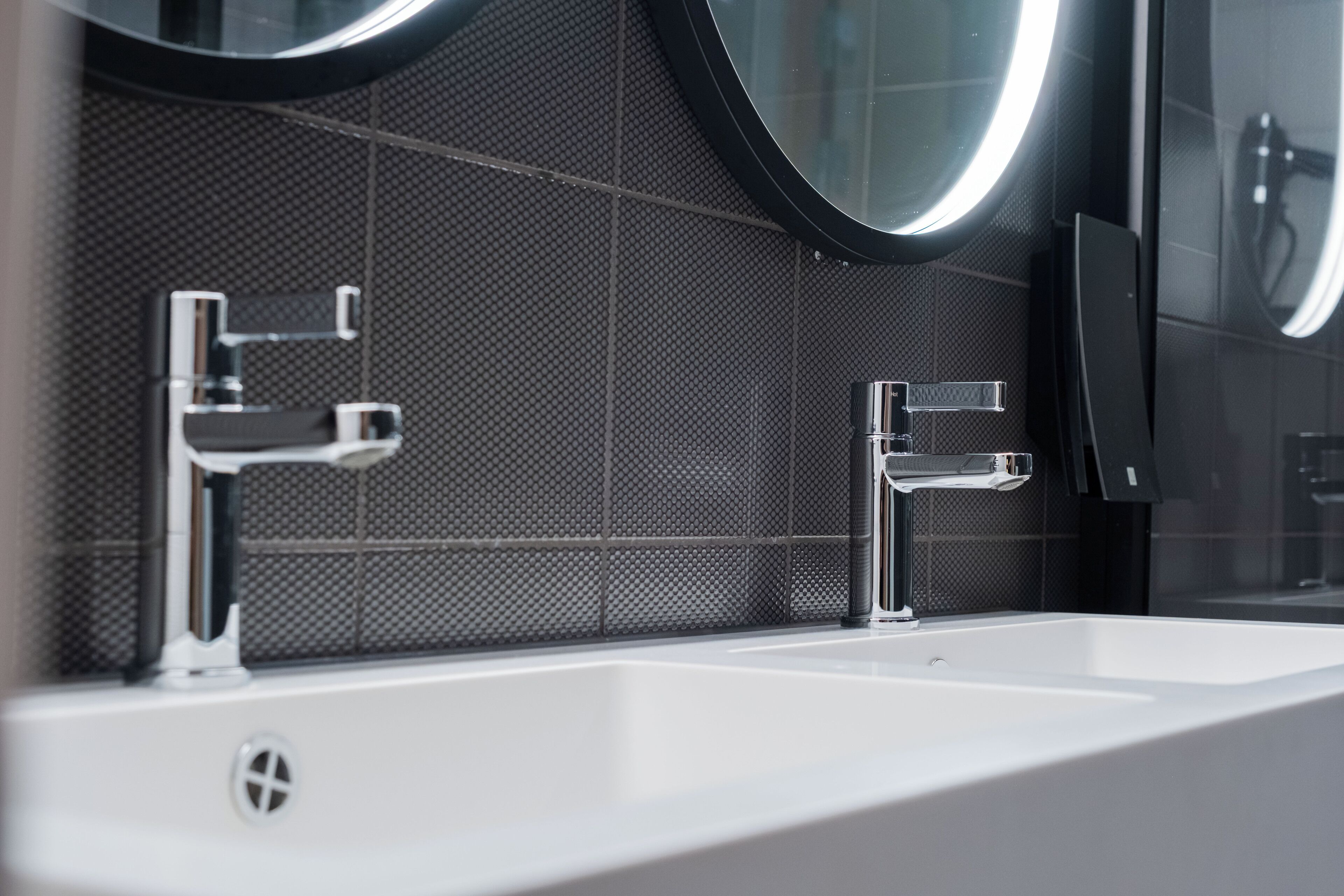 A modern bathroom with a white sink, two chrome faucets, and a large mirror with a circular frame.