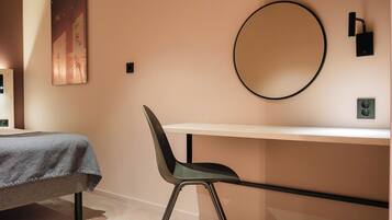 A room with a bed, a desk, a chair, a round mirror, and a wall-mounted light fixture.