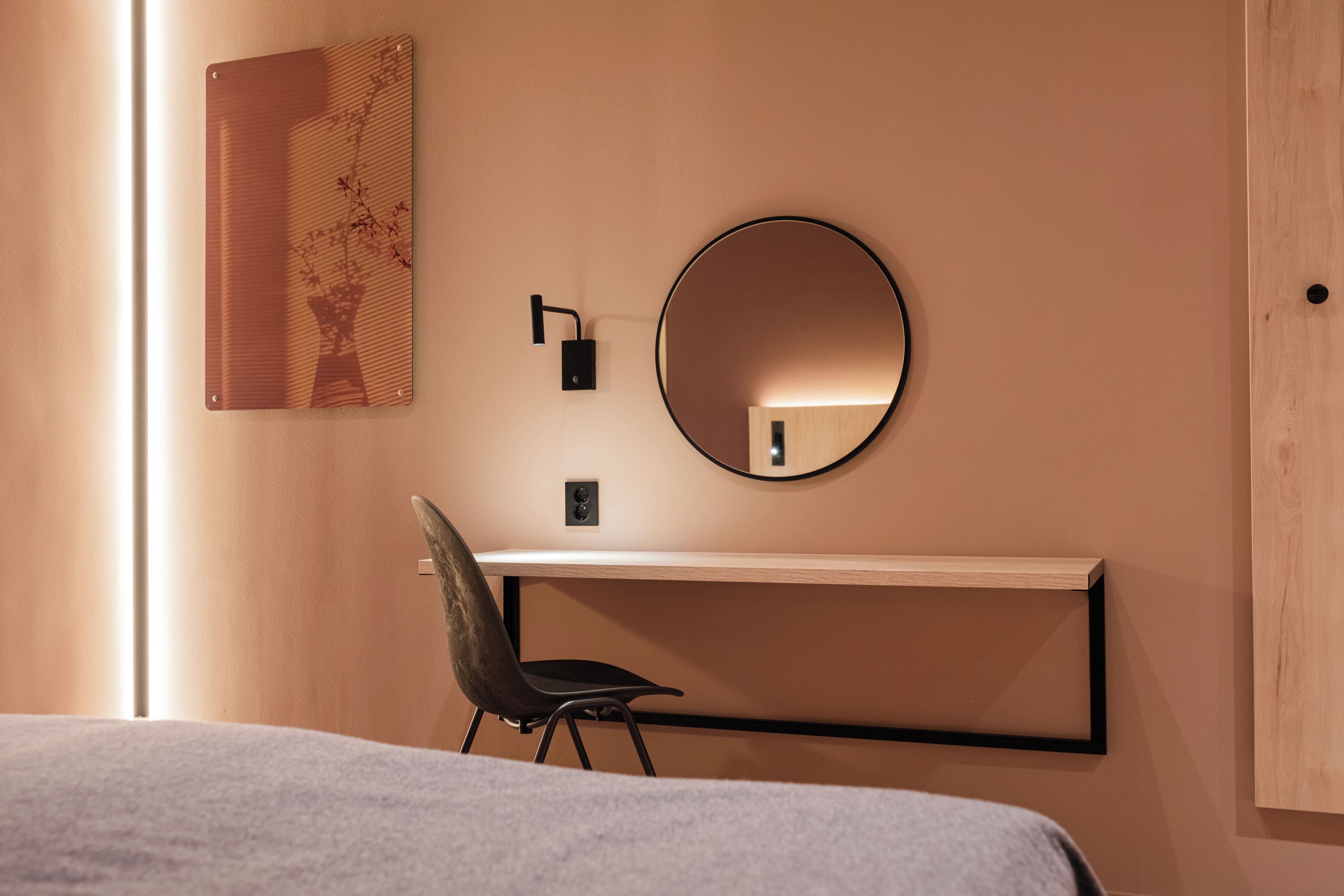 Una habitación de hotel con cama, un escritorio con silla, un espejo redondo y un aplique de pared.