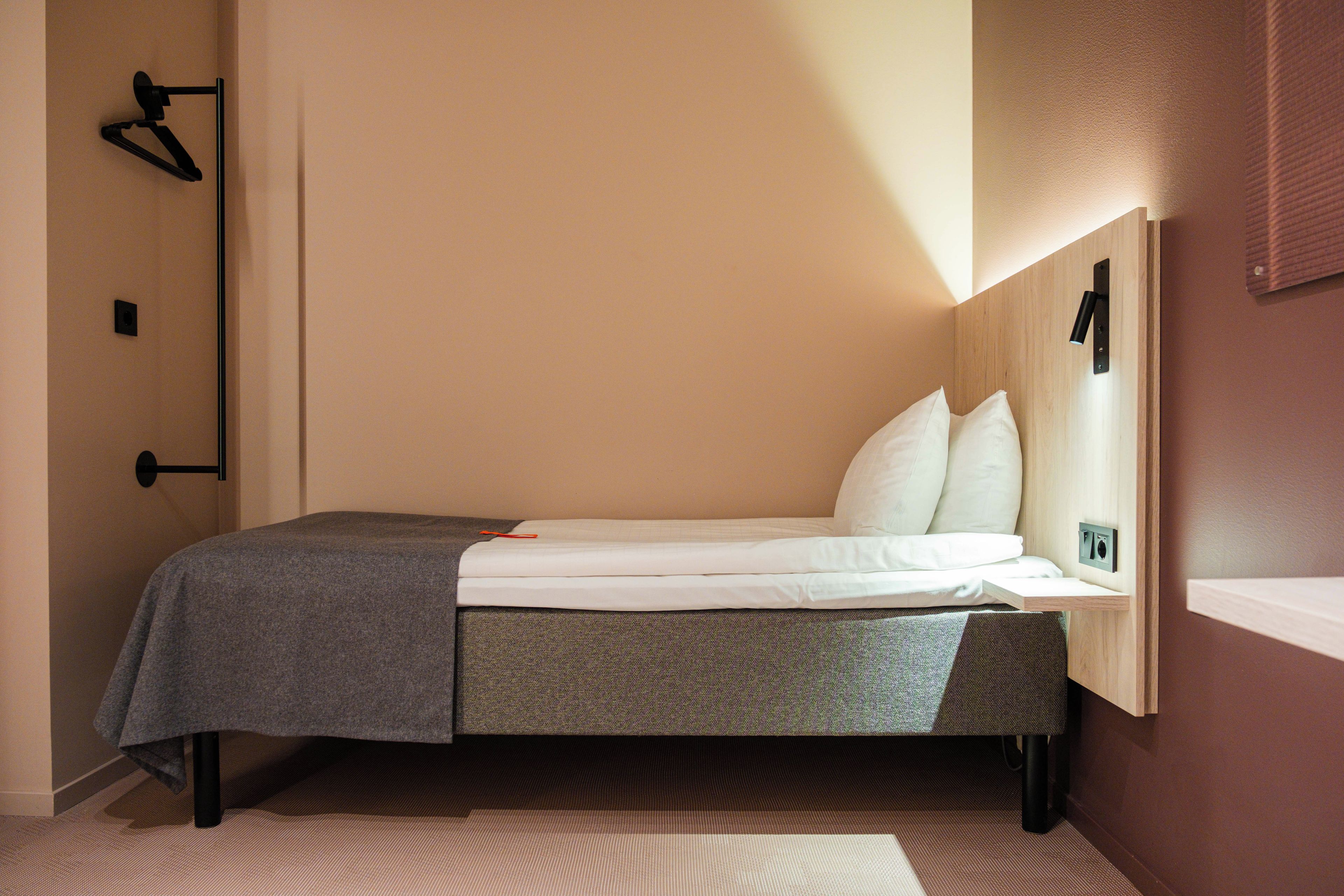 Una cama individual con una manta gris y almohadas blancas en una habitación con cabecera de madera y un aplique de pared.
