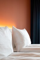 A neatly made bed with white linens and pillows, a plain orange wall, and a dark curtain to the right.